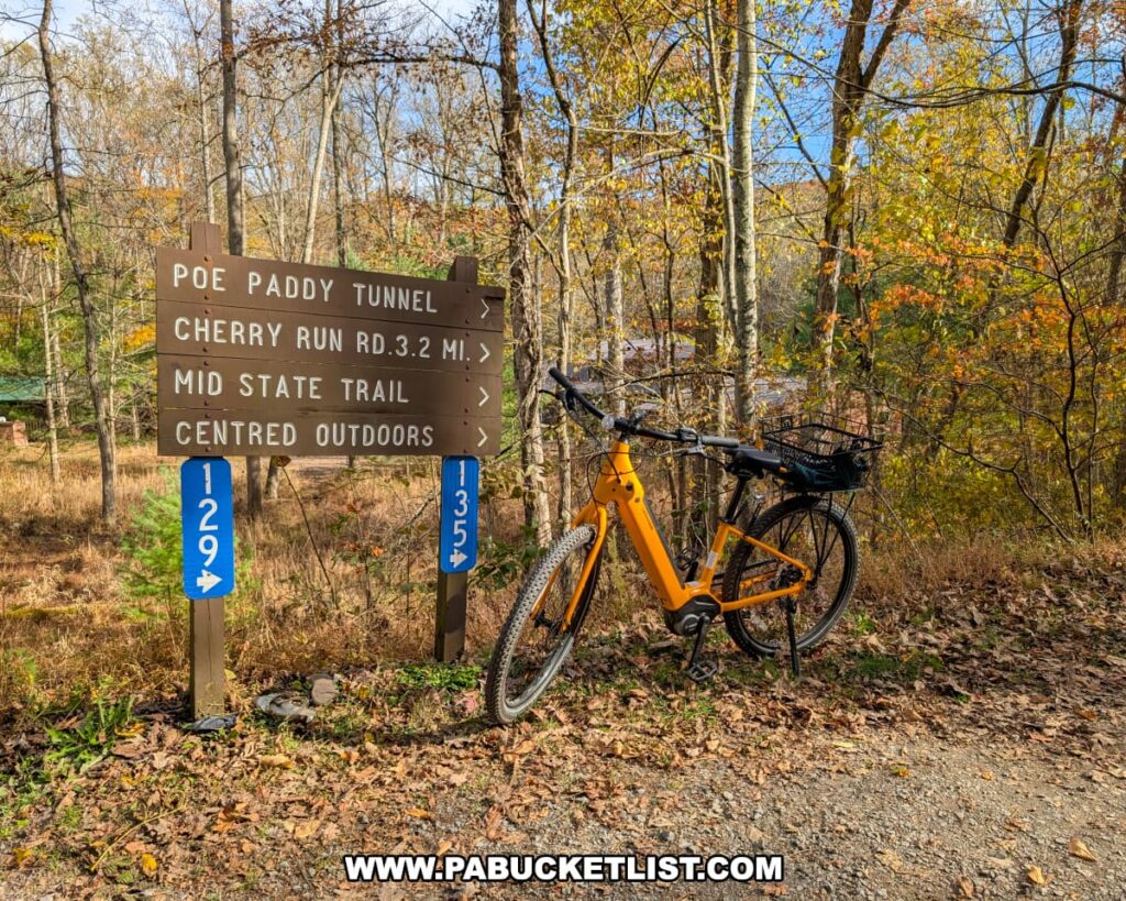 An orange bicycle with a black basket is parked beside a wooden trail sign at the western terminus of the Penn’s Creek Path in Bald Eagle State Forest, surrounded by autumn trees and golden foliage under a clear blue sky.