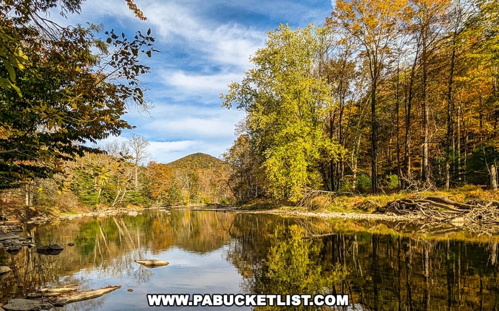 Peaceful autumn scene along Penns Creek at Poe Paddy State Park in Centre County, with golden and green trees reflecting on the calm water and a forested hill rising in the distance beneath a partly cloudy blue sky.