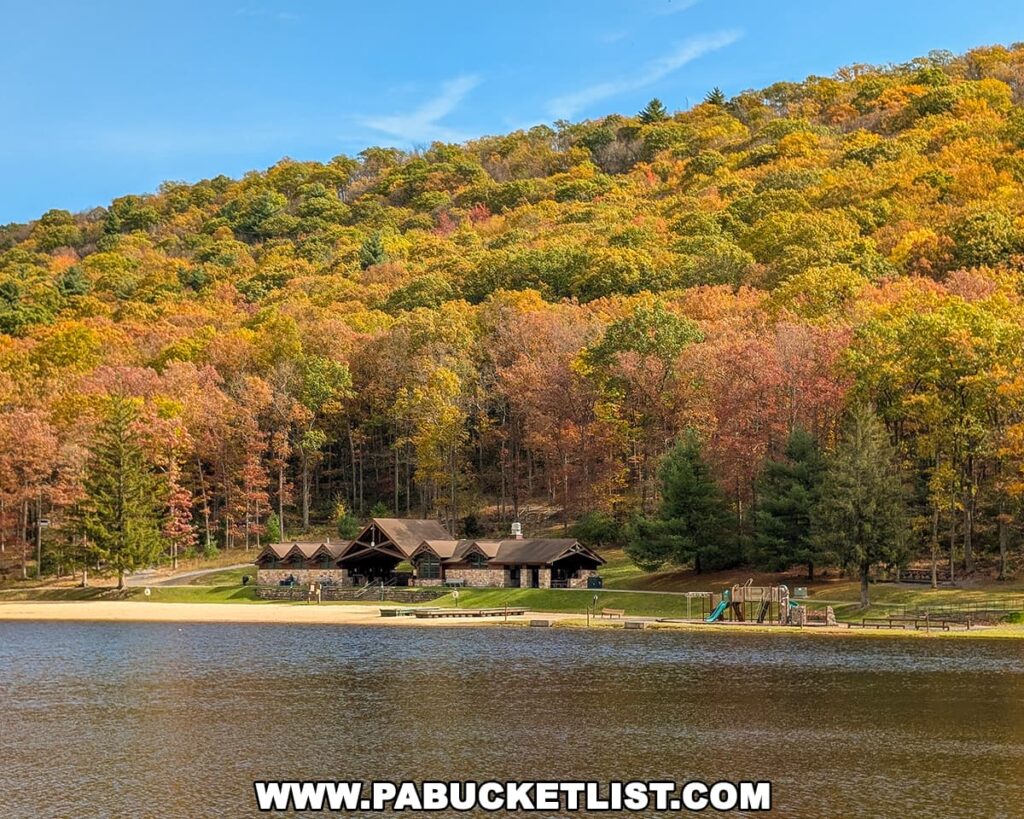 View across Poe Lake at Poe Valley State Park in Centre County, showing the sandy beach, lakeside lodge, and playground nestled below a hillside covered in vibrant fall foliage beneath a bright blue sky.
