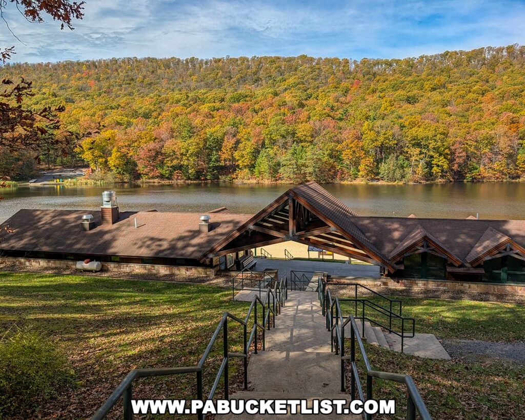 View looking down a long set of concrete steps toward the rustic lakeside lodge and beach area at Poe Valley State Park in Centre County, surrounded by forested hills blanketed in vibrant fall foliage reflected in the calm waters of Poe Lake.