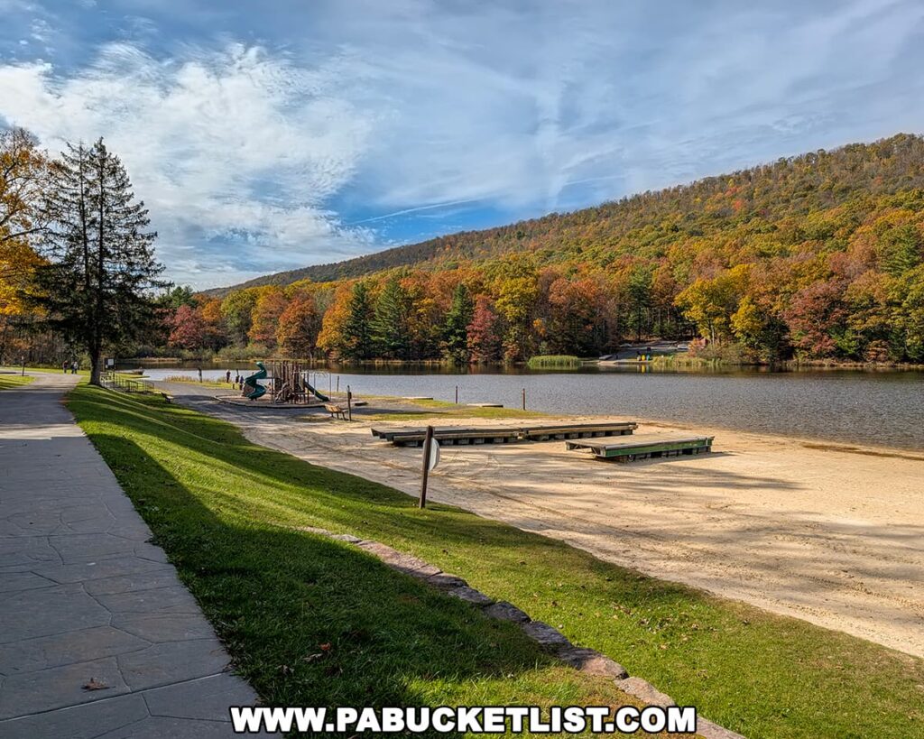 Sandy beach and boat launch area at Poe Valley State Park in Centre County, bordered by a playground and walking path, with forested hillsides glowing in autumn colors reflected on the calm surface of Poe Lake under a partly cloudy sky.