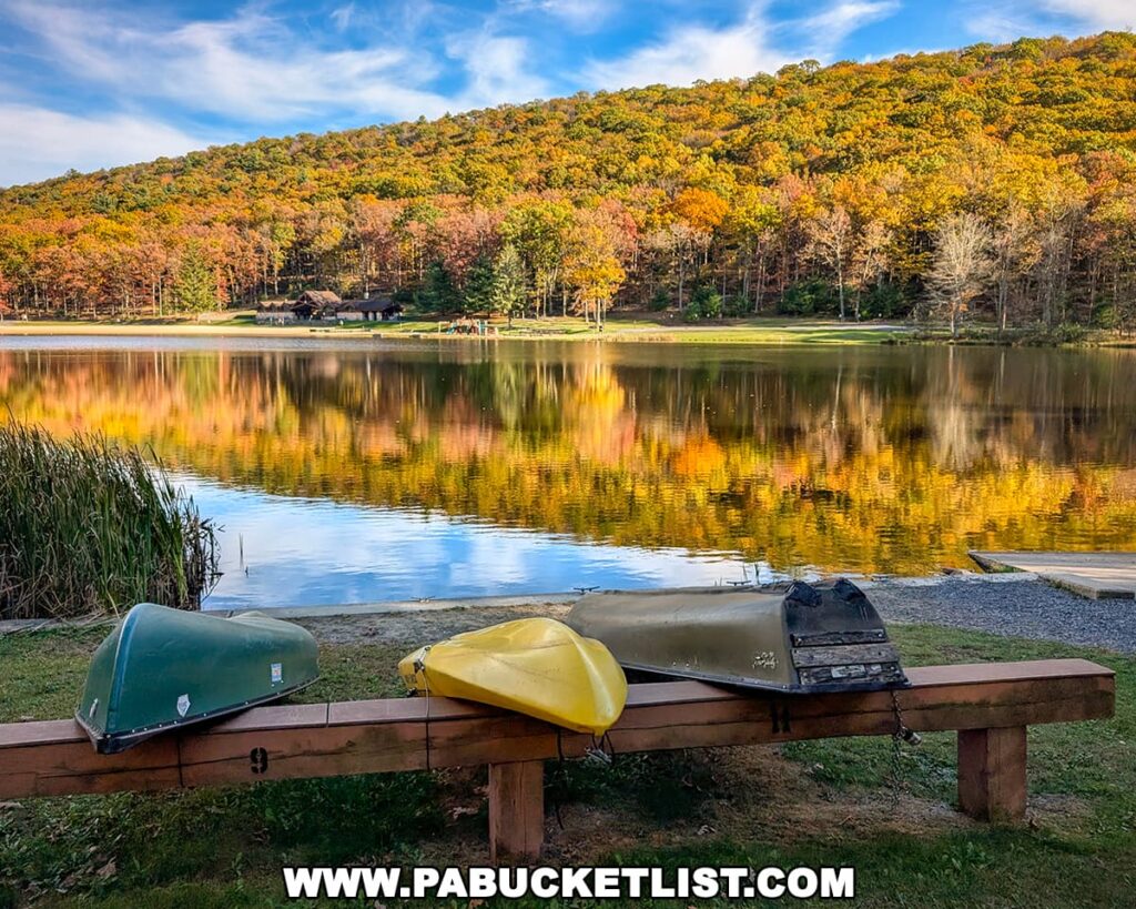 Colorful canoes rest upside down on a wooden bench along the shore of Poe Lake at Poe Valley State Park in Centre County, with vibrant autumn foliage and the lakeside lodge beautifully reflected in the calm, mirror-like water under a bright blue sky.