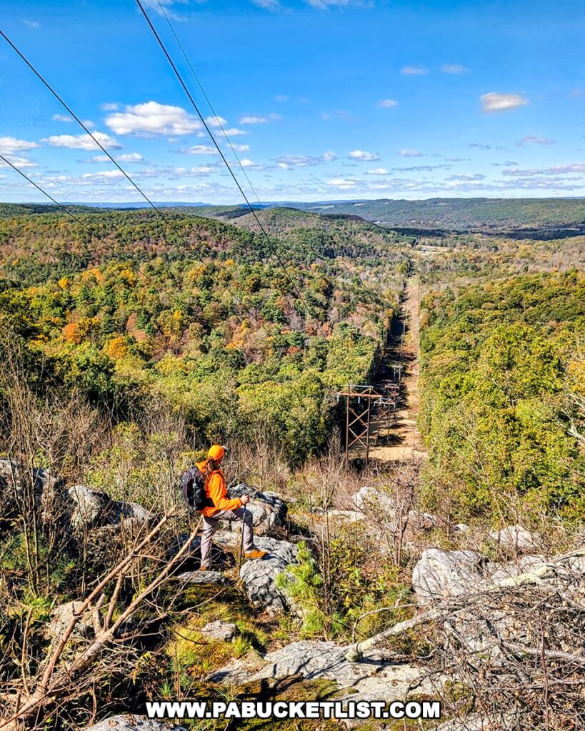 A hiker in an orange jacket stands on a rocky overlook near the Three Sisters Rock Formation, gazing out across a forested valley and powerline corridor stretching toward distant ridges under a bright blue sky in the Rocky Ridge Natural Area of Huntingdon County, Pennsylvania.