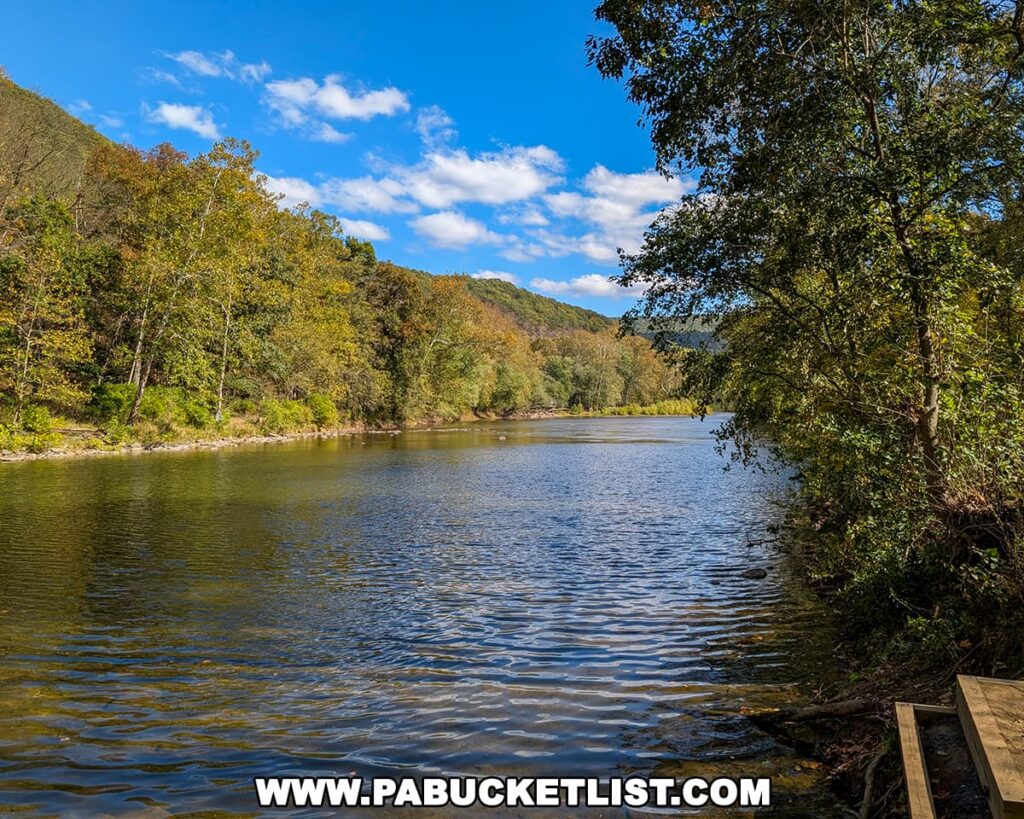 The Raystown Branch of the Juniata River flows gently through Warriors Path State Park in Bedford County, Pennsylvania, surrounded by lush green and early autumn trees beneath a bright blue sky dotted with white clouds.
