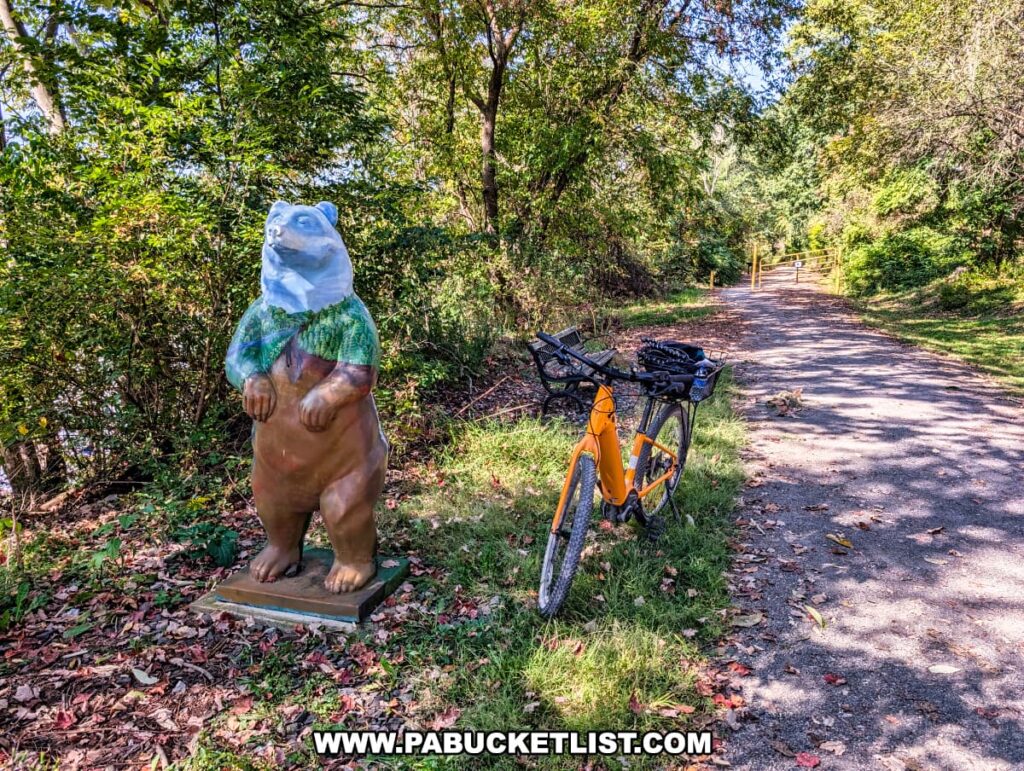 Colorful bear statue dressed in a painted woodland scene standing beside an orange bicycle along the shaded Redbank Valley Rail Trail in western Pennsylvania.