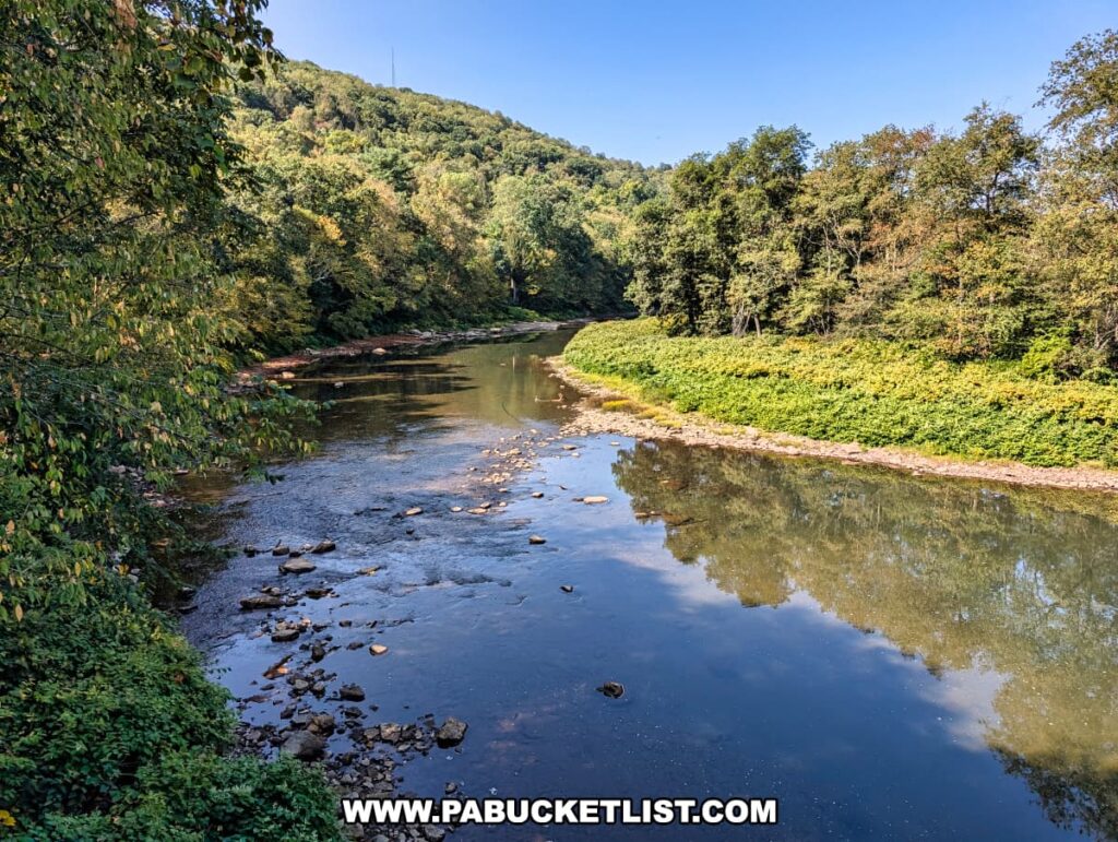 View of Redbank Creek near South Bethlehem along the Redbank Valley Rail Trail, with calm reflective water bordered by rocky shallows, dense forest, and rolling green hills under a clear blue sky.