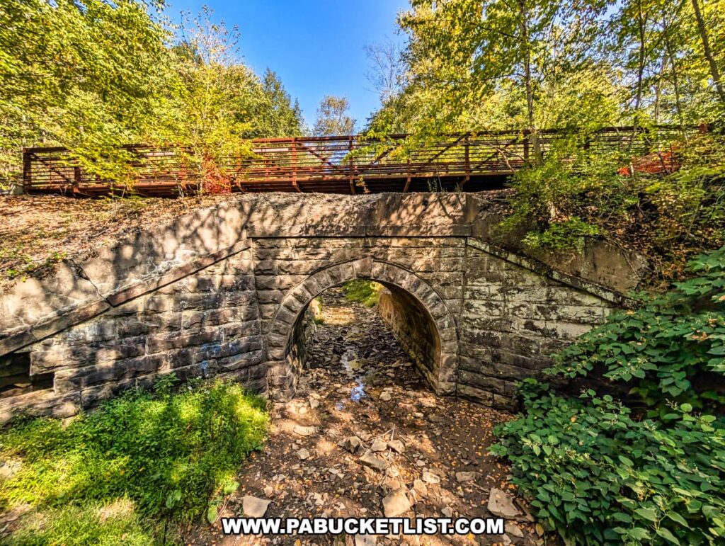 Stone arch foundation of the Terry Mateer Bridge along the Redbank Valley Rail Trail, with the modern steel and wood trail bridge crossing above a rocky streambed and surrounded by dense forest.