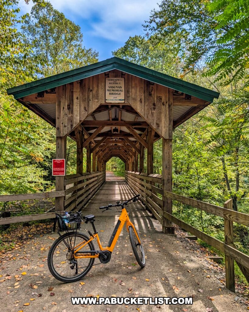 Orange bicycle parked in front of the Geraldine L. and Richard F. Vidunas Memorial Bridge, a covered wooden bridge surrounded by lush green forest along the Redbank Valley Rail Trail.