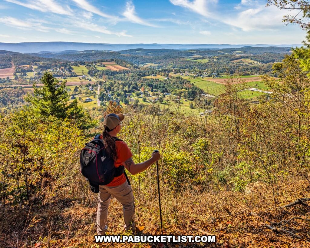 Hiker with a backpack and trekking pole standing at Katy Jane West Vista in Rider Park, Lycoming County, Pennsylvania, overlooking a patchwork of farmland, forested hills, and distant mountain ridges under a clear blue autumn sky.