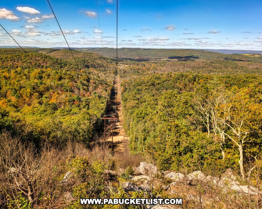 A panoramic autumn view from a rocky overlook near the Three Sisters Rock Formation in the Rocky Ridge Natural Area, showing a powerline corridor cutting through rolling forested ridges under a bright blue sky in Huntingdon County, Pennsylvania.