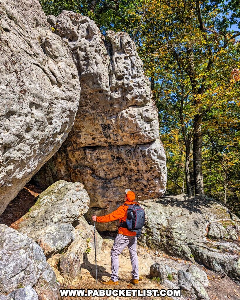 A hiker in an orange jacket stands beneath a massive weathered limestone boulder near the Three Sisters Rock Formation in the Rocky Ridge Natural Area, surrounded by rugged rocks and colorful autumn trees in Huntingdon County, Pennsylvania.