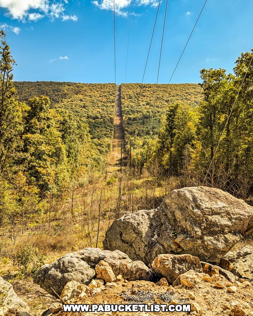A view from a rocky clearing near the Three Sisters Rock Formation in the Rocky Ridge Natural Area, looking southeast along a powerline corridor that cuts a straight path over a forested ridge beneath a bright blue sky in Huntingdon County, Pennsylvania.