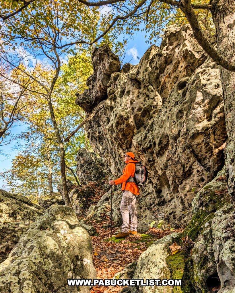A hiker in an orange jacket stands along a rocky trail beneath towering weathered limestone cliffs near the Three Sisters Rock Formation in the Rocky Ridge Natural Area, surrounded by moss-covered boulders and colorful autumn foliage in Huntingdon County, Pennsylvania.