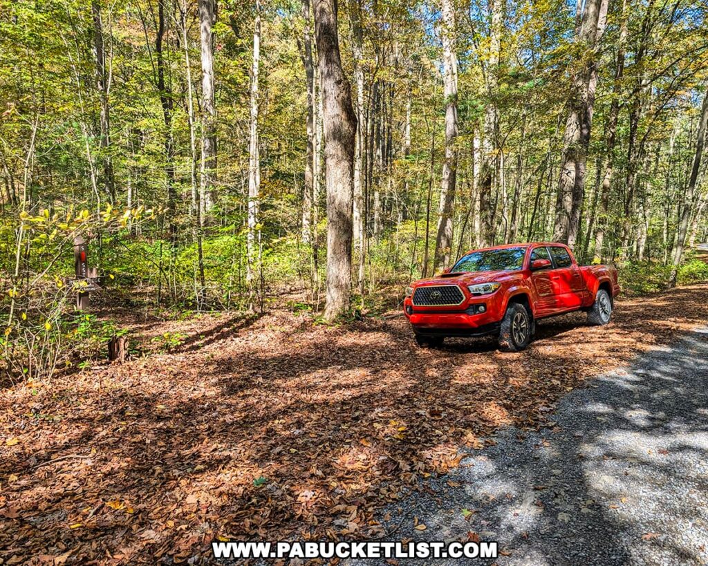 A red pickup truck parked along a leaf-covered forest road near the trailhead for the Three Sisters Rock Formation in the Rocky Ridge Natural Area, surrounded by tall trees and early autumn foliage in Huntingdon County, Pennsylvania.