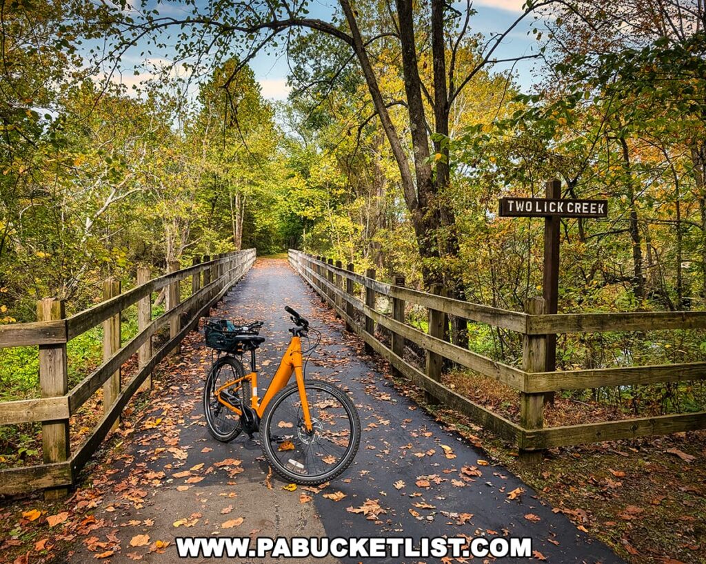 A bright yellow bicycle is parked on a wooden bridge crossing Two Lick Creek along the Hoodlebug Trail in Indiana County, Pennsylvania. Autumn leaves are scattered across the paved surface, and a wooden sign marks the creek’s name. The bridge is surrounded by colorful fall foliage under a partly cloudy sky, creating a peaceful, scenic moment along the 10-mile rail trail that follows the former Pennsylvania Railroad’s Indiana Branch, once traveled by the self-propelled “Hoodlebug” passenger coach.