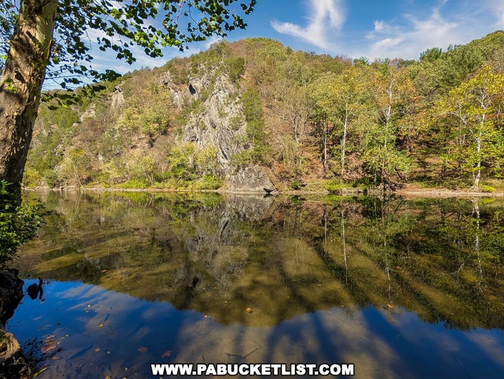 Cliffs and early autumn trees reflect in the calm, clear waters of the Raystown Branch of the Juniata River at Warriors Path State Park in Bedford County, Pennsylvania, beneath a bright blue sky with wispy clouds.