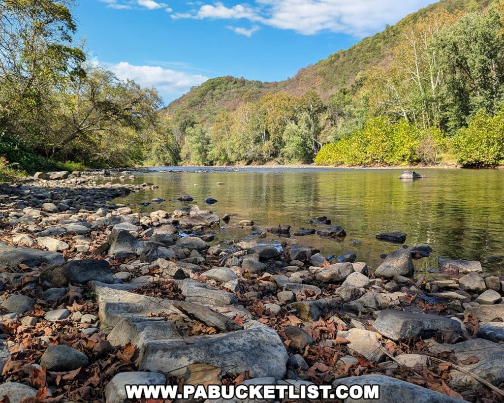 A rocky shoreline along the Raystown Branch of the Juniata River at Warriors Path State Park in Bedford County, Pennsylvania, with tree-covered hills rising in the distance under a bright blue sky.