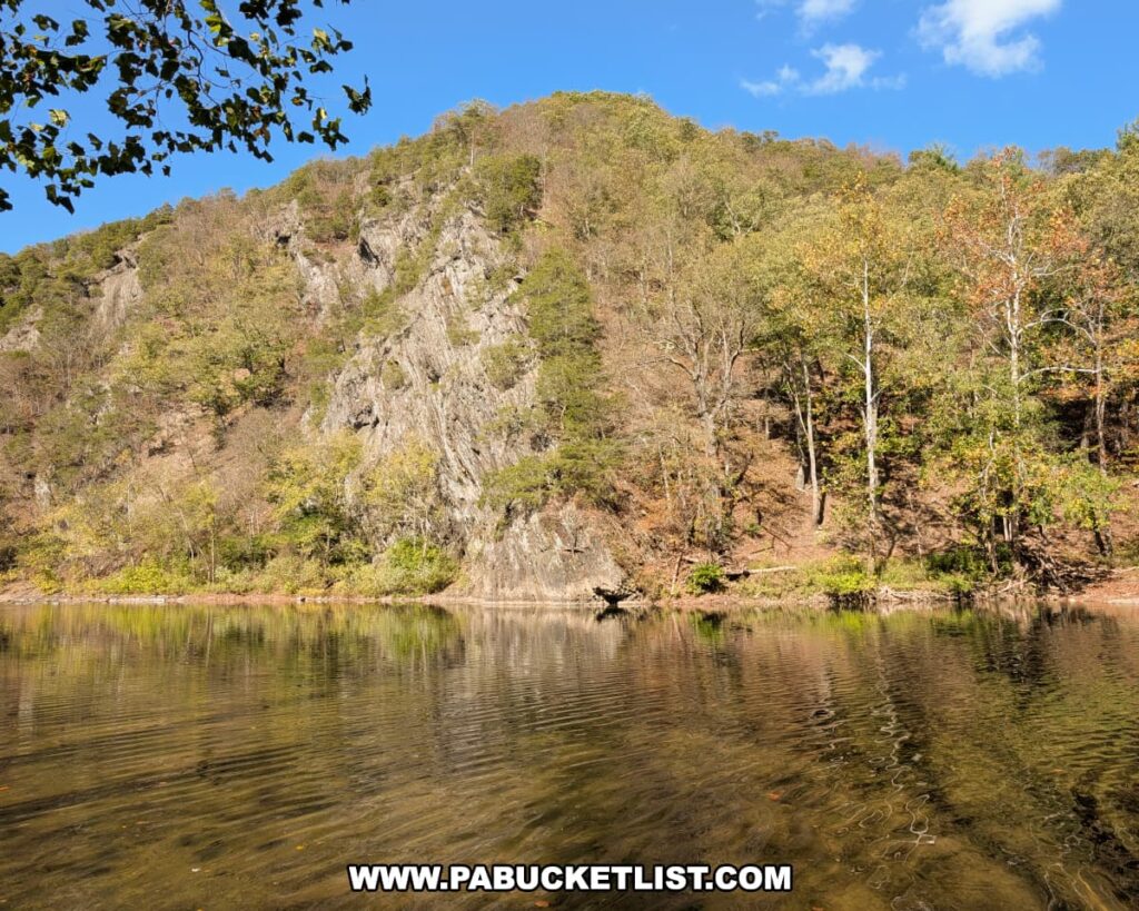 Steep rocky cliffs rise above the calm, reflective waters of the Raystown Branch of the Juniata River at Warriors Path State Park in Bedford County, Pennsylvania, surrounded by early autumn foliage under a clear blue sky.