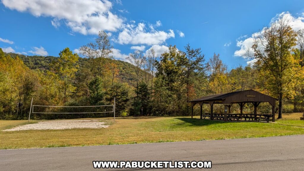 A sand volleyball court and wooden picnic pavilion sit on a grassy field surrounded by wooded hills with early autumn foliage at Warriors Path State Park in Bedford County, Pennsylvania, under a bright blue sky with scattered clouds.