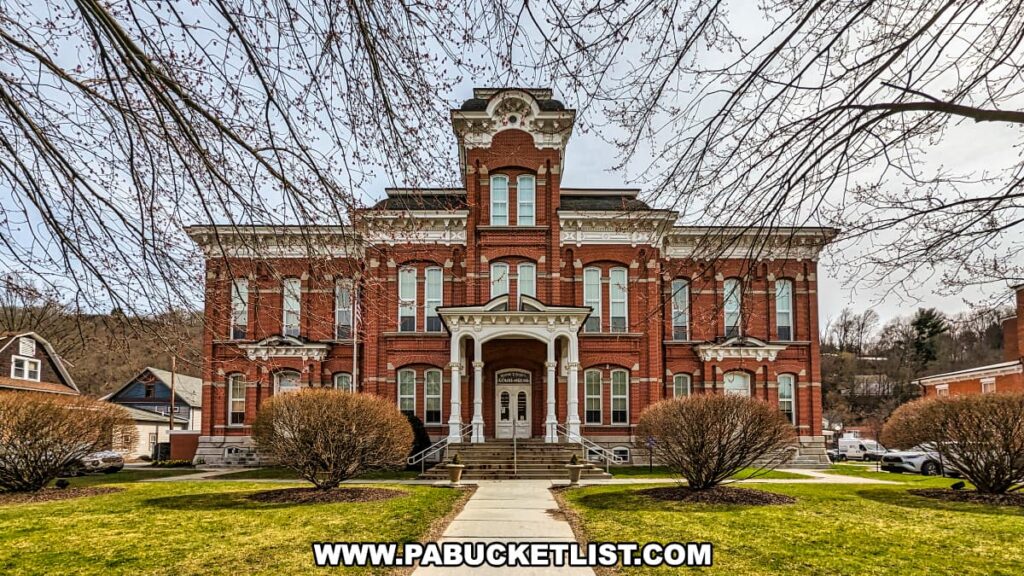 Front view of the historic Wayne County Courthouse in Honesdale, Pennsylvania, featuring a symmetrical red-brick façade with tall arched windows, white decorative trim, and a central tower rising above the main entrance. The courthouse is framed by bare tree branches and surrounded by neatly trimmed shrubs and a manicured lawn under an overcast sky.