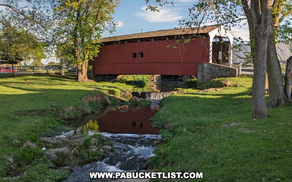 Willows Covered Bridge in Lancaster County, Pennsylvania, viewed from downstream with evening sunlight illuminating its red wooden sides, reflecting in a small creek that flows beneath the bridge surrounded by lush green grass and mature trees.
