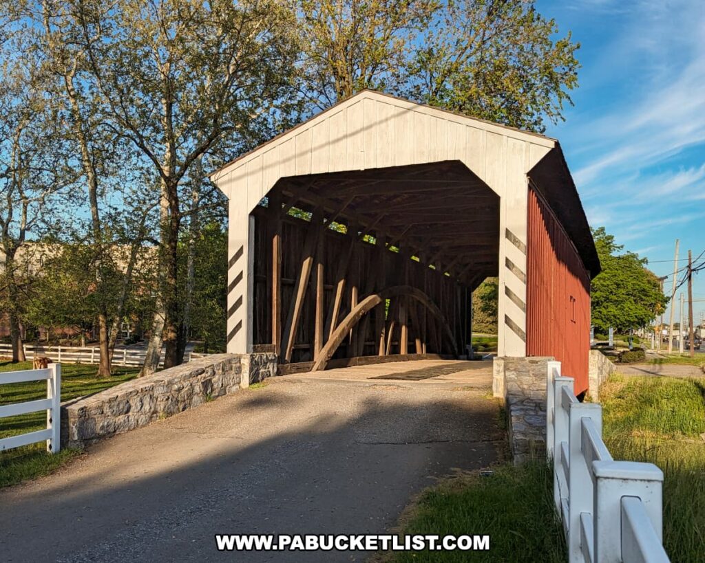 Front view of the Willows Covered Bridge in Lancaster County, Pennsylvania, showing its white portal, red wooden sides, and exposed timber arches under a bright blue sky, framed by stone walls and white fencing along the entrance road.