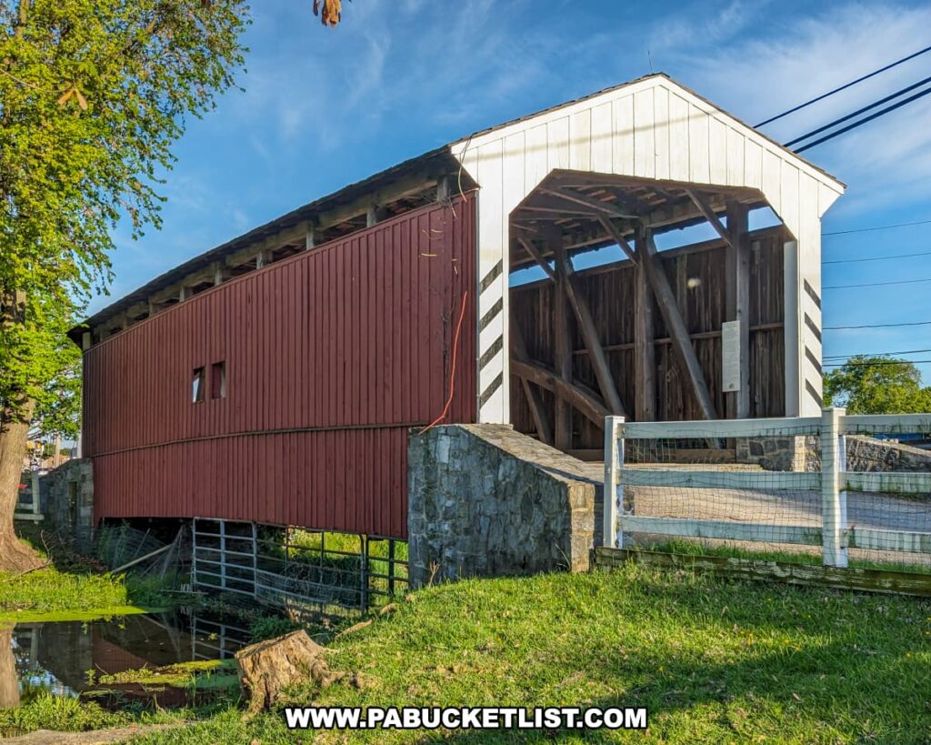 Side view of the Willows Covered Bridge in Lancaster County, Pennsylvania, showing its red wooden walls, white portal with diagonal stripes, stone abutments, and a small stream reflecting the bridge beneath a bright blue sky.