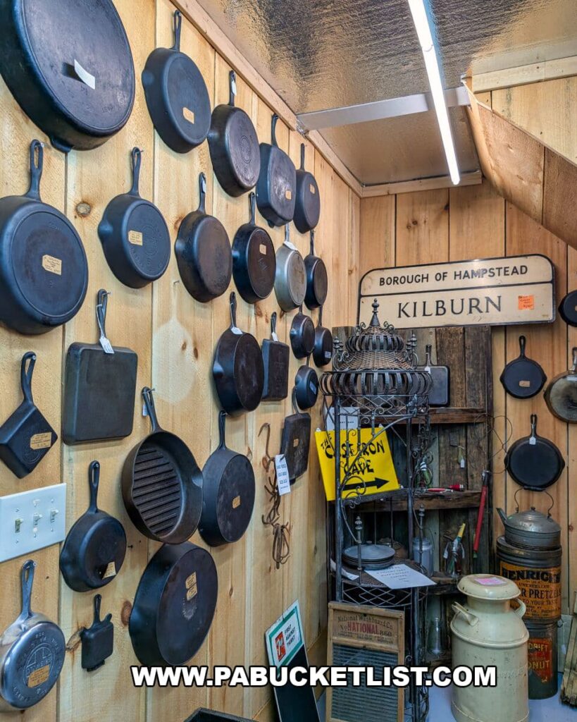 Wall display of vintage cast-iron skillets and cookware at Apple Hill Antiques near State College, with additional antique signs, kitchen tools, and rustic collectibles arranged throughout the corner of a wood-paneled room.