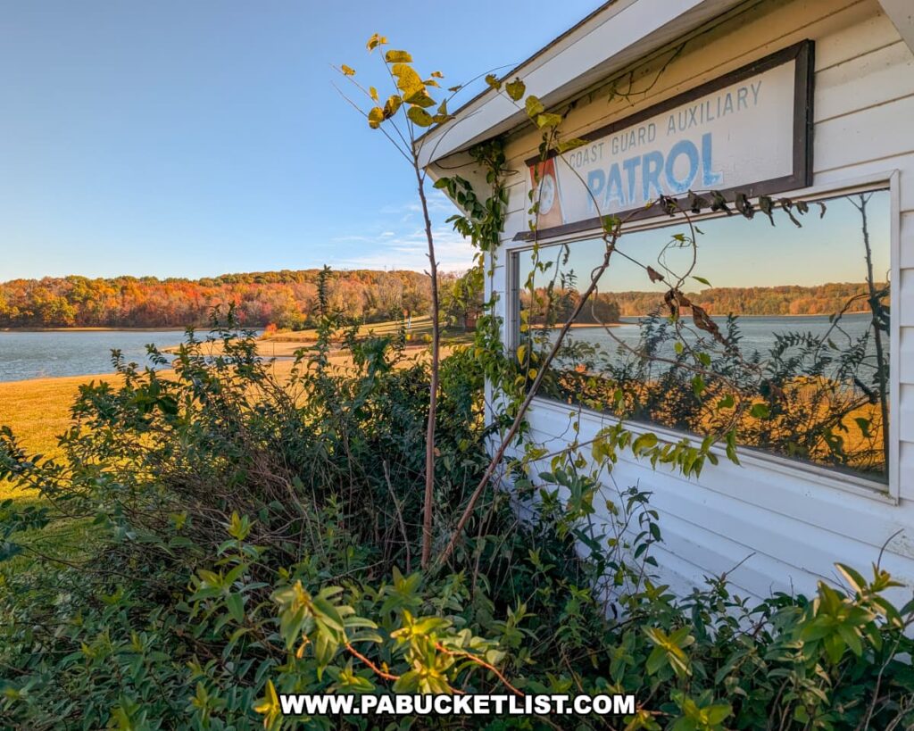 An old Coast Guard Auxiliary Patrol building partially covered in vines overlooks Lake Marburg at Codorus State Park in York County, Pennsylvania, with colorful autumn foliage reflecting in the window and bright sunlight illuminating the scene.