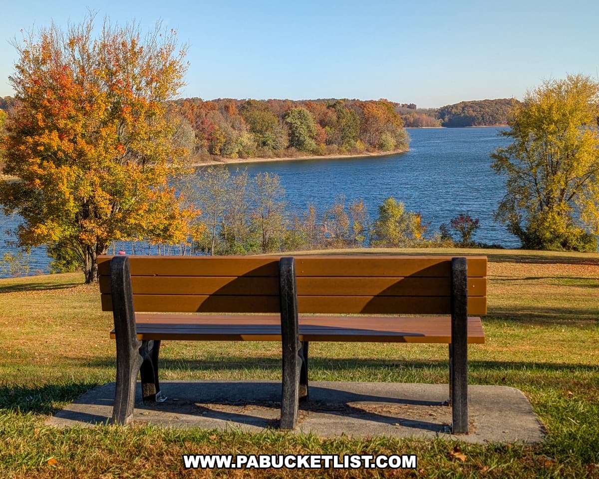 A wooden bench overlooks the calm blue waters of Lake Marburg at Codorus State Park in York County, Pennsylvania, surrounded by trees glowing with autumn colors under a clear blue sky.