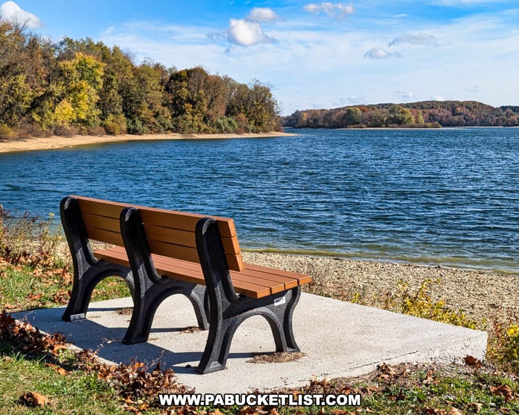 Two benches on a concrete pad overlook the deep blue waters of Lake Marburg at Codorus State Park in York County, Pennsylvania, with wooded hills displaying late autumn colors in the distance under a partly cloudy sky.