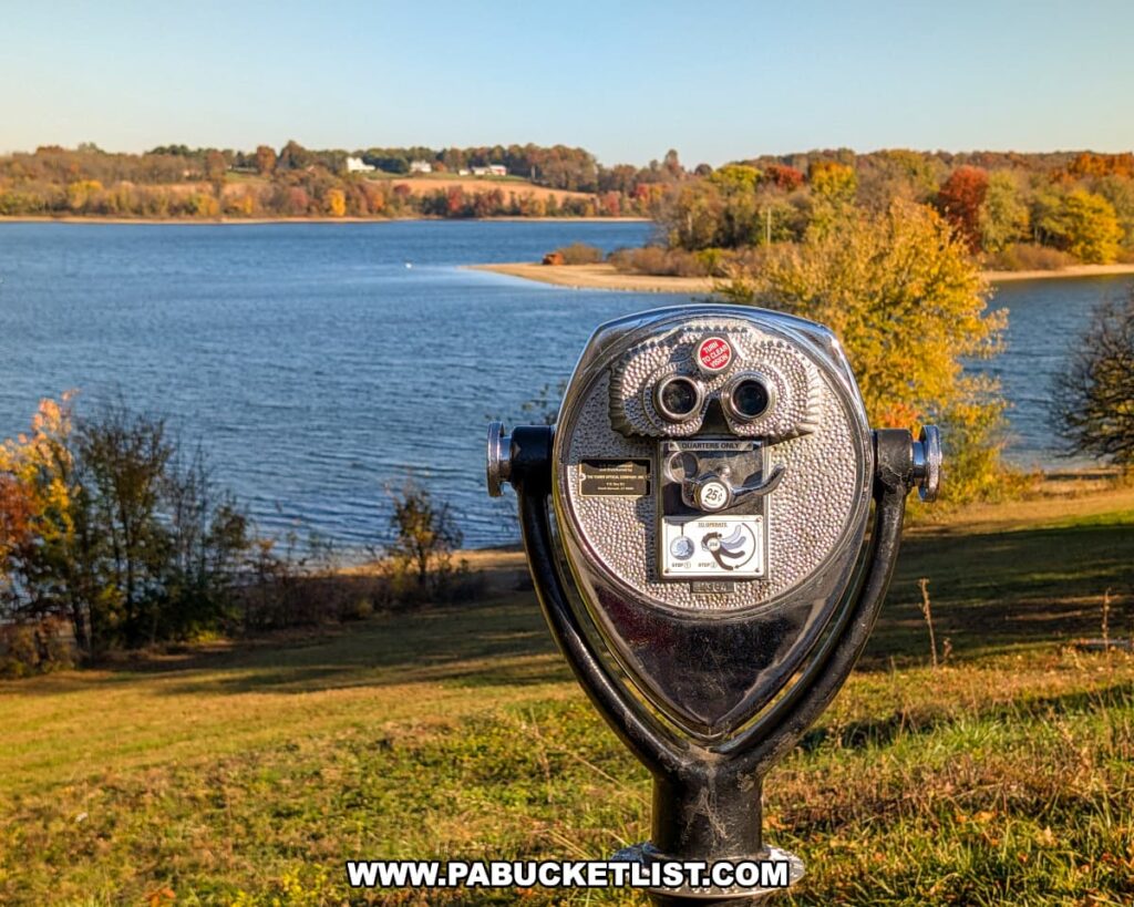 A coin-operated binocular viewer overlooks the blue waters of Lake Marburg at Codorus State Park in York County, Pennsylvania, with colorful autumn trees and rolling hills visible in the distance under a clear sky.