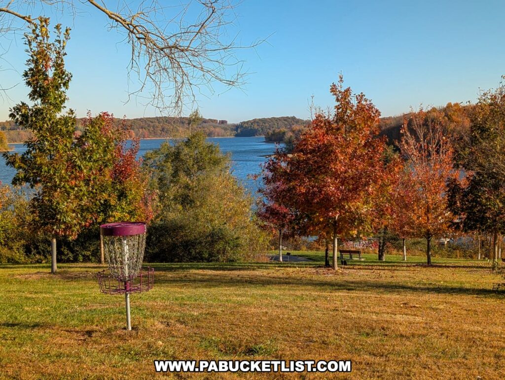 A disc golf basket stands on a grassy hill overlooking the blue waters of Lake Marburg at Codorus State Park in York County, Pennsylvania, surrounded by colorful autumn trees under a clear blue sky.