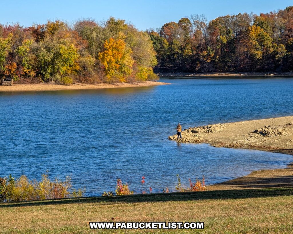 A lone fisherman stands on a rocky shoreline along the calm blue waters of Lake Marburg at Codorus State Park in York County, Pennsylvania, surrounded by colorful autumn trees under a clear sky.