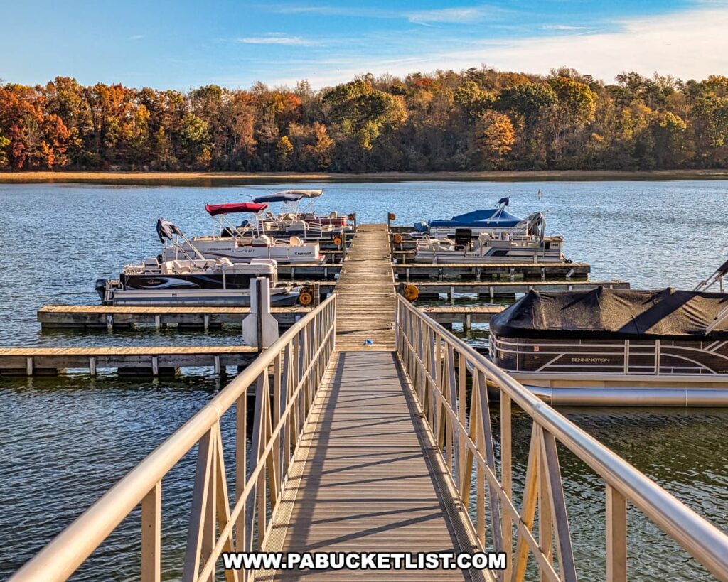 A wooden dock lined with pontoon boats extends into the calm blue waters of Lake Marburg at Codorus State Park in York County, Pennsylvania, with autumn-colored trees reflecting in the distance under a soft afternoon sky.