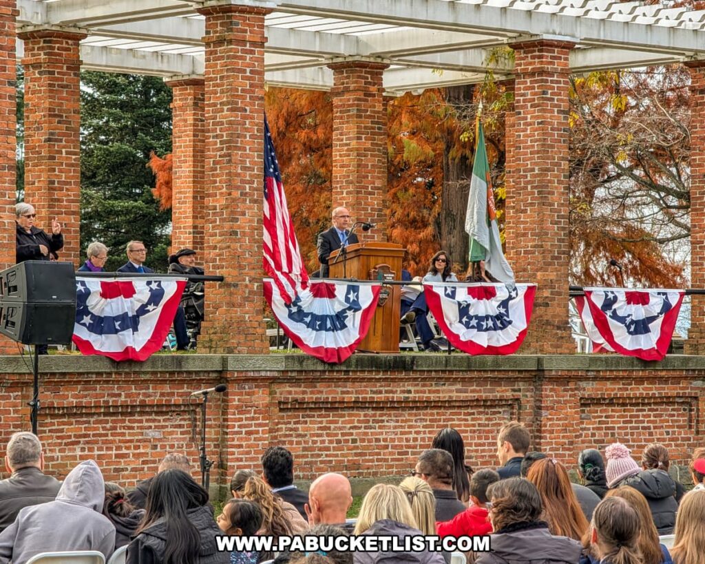 A visiting dignitary speaks from a podium decorated with patriotic bunting under a brick pavilion at Soldiers’ National Cemetery in Gettysburg during the Dedication Day ceremony as the audience listens attentively.