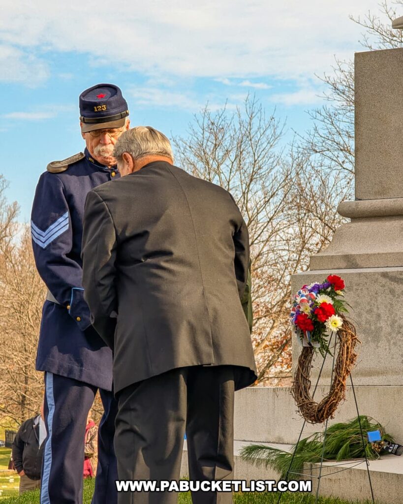 A Civil War reenactor and another participant bow their heads beside a monument adorned with a wreath of red, white, and blue flowers during the wreath-laying portion of the Dedication Day ceremony at Soldiers’ National Cemetery in Gettysburg.