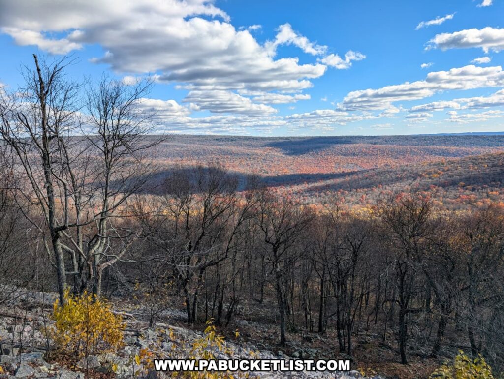 Expansive view from Hubler Gap Vista in Rothrock State Forest near State College, showing a rocky foreground and leafless trees overlooking rolling ridges covered in late autumn foliage under a blue sky with scattered clouds.