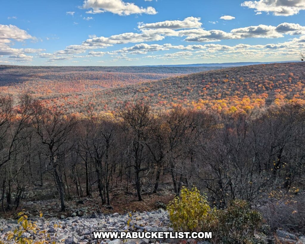 Scenic autumn view from Hubler Gap Vista in Rothrock State Forest near State College, showing a rocky slope and leafless trees overlooking rolling ridges blanketed in orange and gold foliage under a blue sky with scattered clouds.