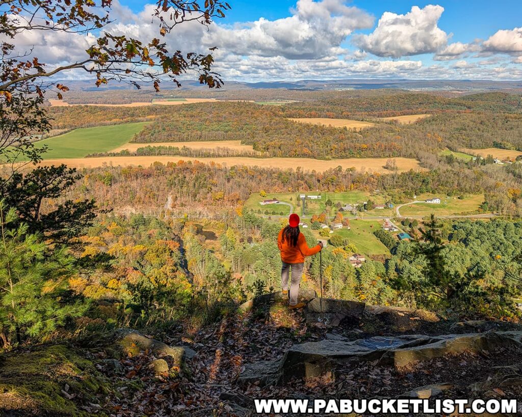 Hiker in an orange jacket standing on a rocky ledge at Indian Lookout in Rothrock State Forest, gazing out over a sweeping patchwork of fields, forests, and rolling ridges glowing with October foliage under a bright, cloud-dotted sky.
