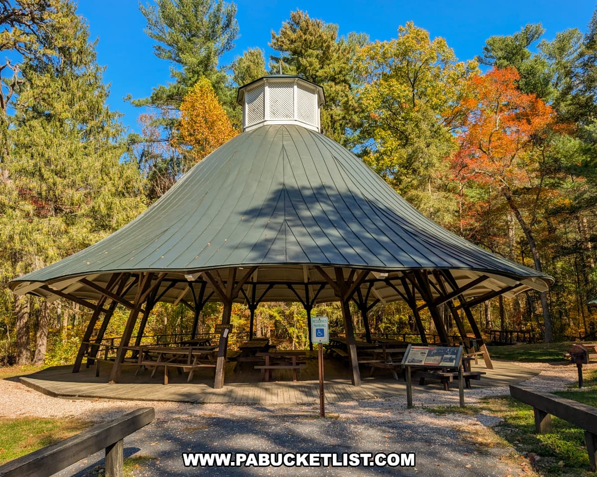 The distinctive round dance pavilion at Mont Alto State Park in Franklin County, Pennsylvania, with its green metal roof, picnic tables, and surrounding trees glowing with vibrant autumn colors under a clear blue sky.