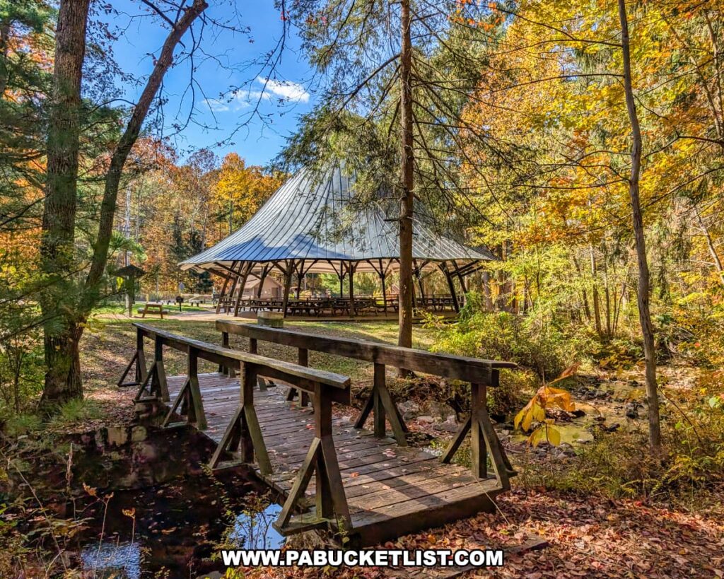 A charming wooden footbridge crossing the West Branch of Antietam Creek at Mont Alto State Park in Franklin County, Pennsylvania, with the park’s distinctive round dance pavilion and colorful autumn foliage in the background.