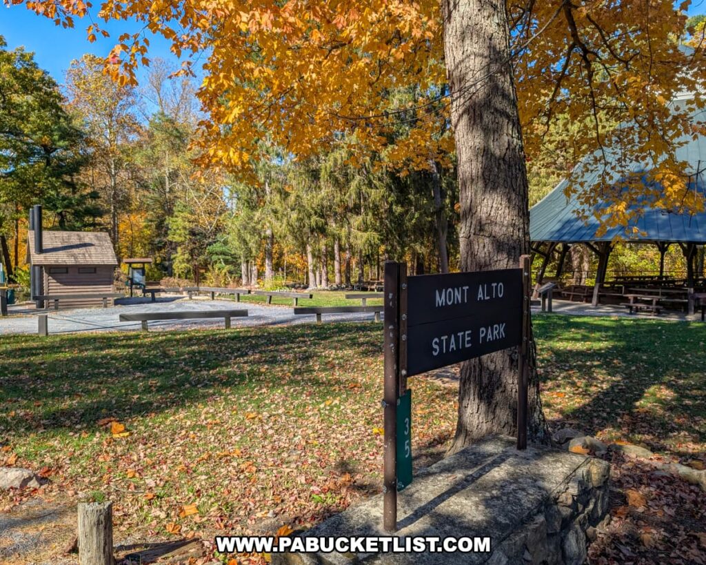 A Mont Alto State Park sign mounted on a stone base beneath golden autumn leaves, with a picnic pavilion and rustic log restroom building visible in the background at Mont Alto State Park in Franklin County, Pennsylvania.