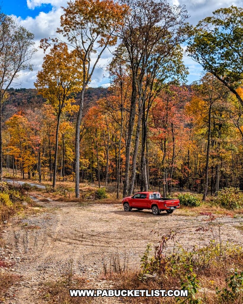 Red pickup truck parked at the Pump Station Trail parking area along Harry’s Valley Road in Rothrock State Forest, surrounded by tall trees showcasing peak autumn colors under a bright blue sky with scattered clouds.