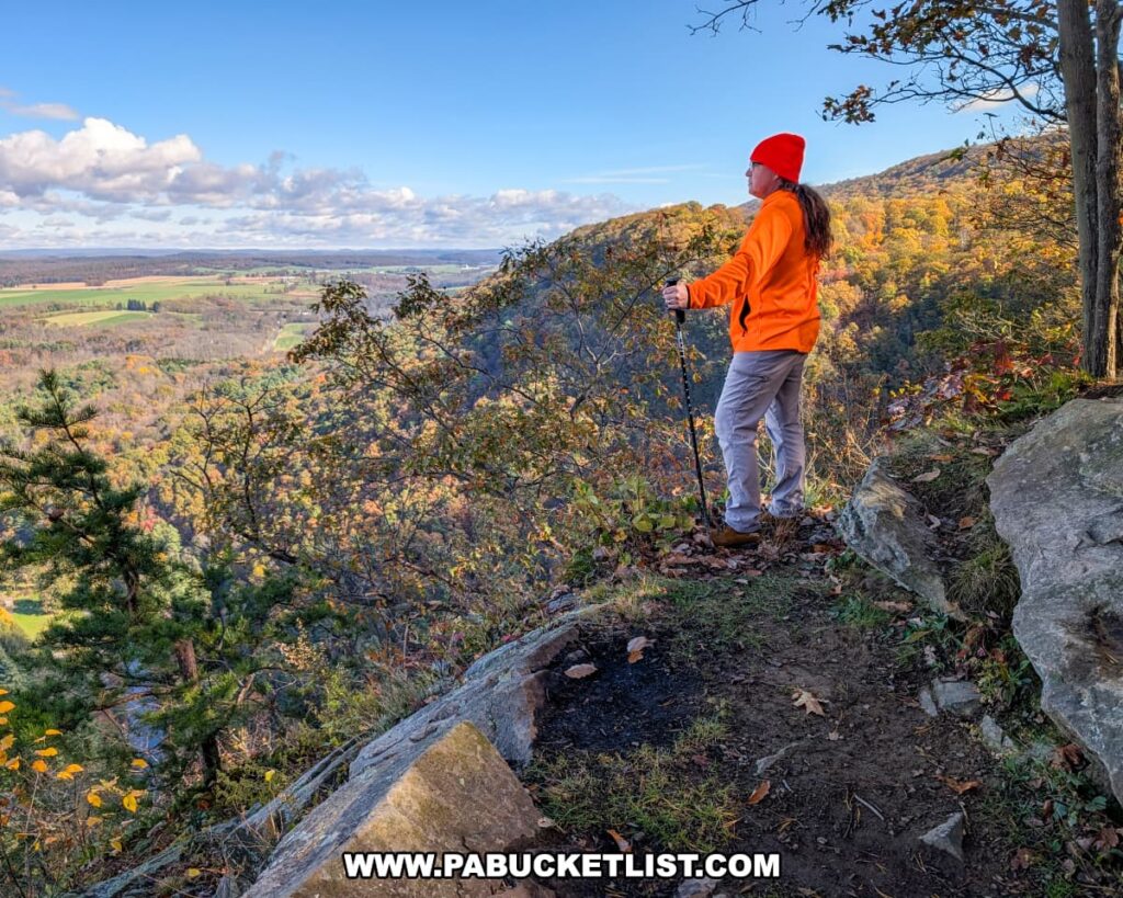 Hiker in an orange jacket and red hat standing on a rocky ledge at Indian Lookout in Rothrock State Forest, overlooking a sweeping valley of colorful October foliage, farmland, and distant ridges under a bright blue sky.