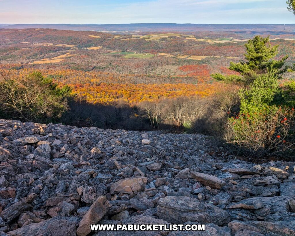 View from Sausser's Stone Pile along the Standing Stone Trail in Huntingdon County, Pennsylvania, featuring a rugged slope of jagged gray rocks overlooking a valley filled with vibrant autumn foliage, rolling hills, and distant farmland under a soft blue sky.