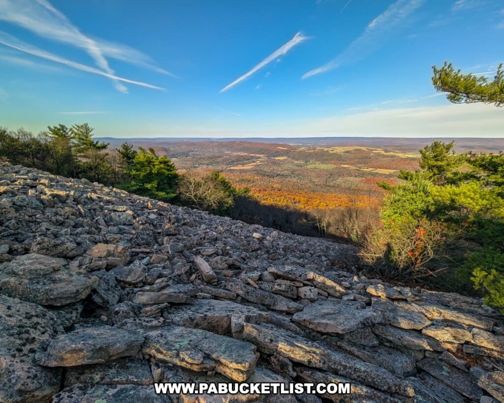 Expansive view from Sausser's Stone Pile along the Standing Stone Trail in Huntingdon County, Pennsylvania, featuring a vast field of angular gray rocks in the foreground and a sweeping vista of forested hills, farmland, and blue sky stretching into the distance.