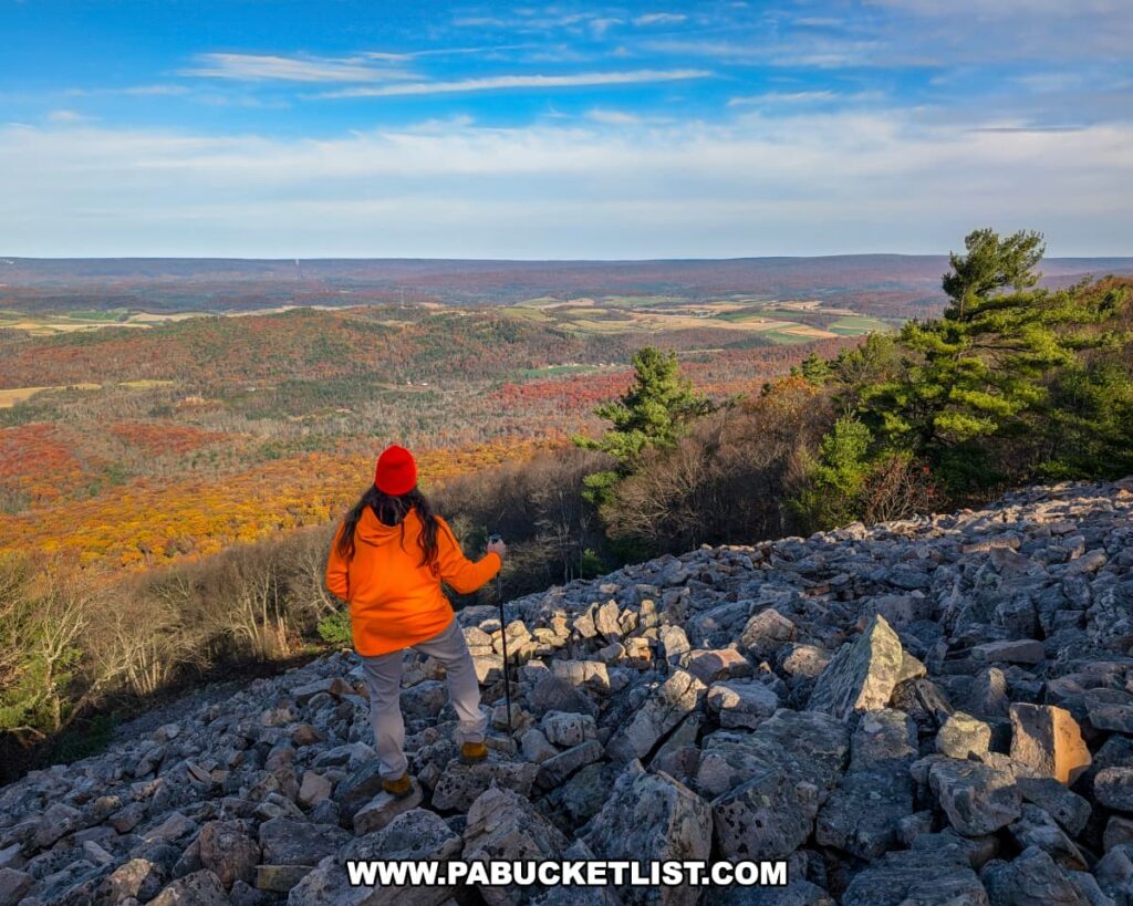A hiker wearing an orange jacket and red hat stands on the rocky expanse of Sausser's Stone Pile along the Standing Stone Trail in Huntingdon County, Pennsylvania, gazing out over a colorful autumn landscape of forests, farmland, and distant ridges beneath a bright blue sky.