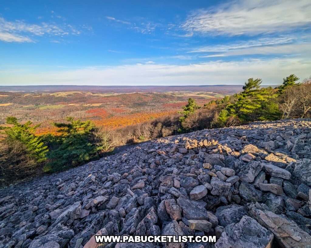 View from Sausser's Stone Pile along the Standing Stone Trail in Huntingdon County, Pennsylvania, showing a vast slope of jagged gray rocks descending toward a colorful valley filled with autumn foliage, evergreen trees, and distant rolling farmland under a clear blue sky.