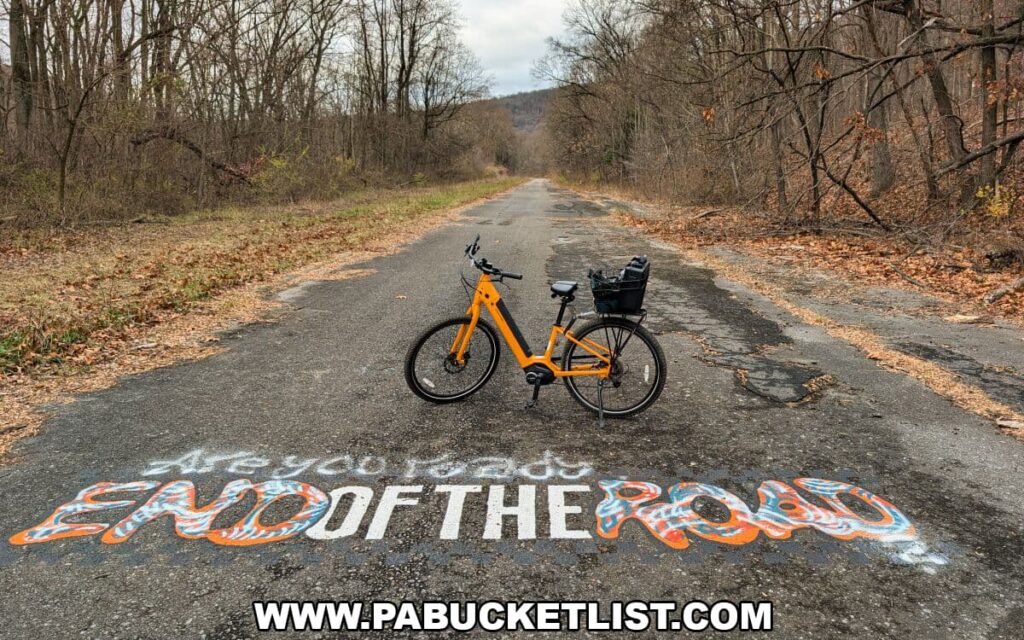 Orange bicycle parked on the cracked, overgrown pavement of the Abandoned PA Turnpike, with colorful “End of the Road” graffiti painted across the old highway and leafless woods lining this 8.5-mile former Turnpike stretch in Bedford and Fulton counties.
